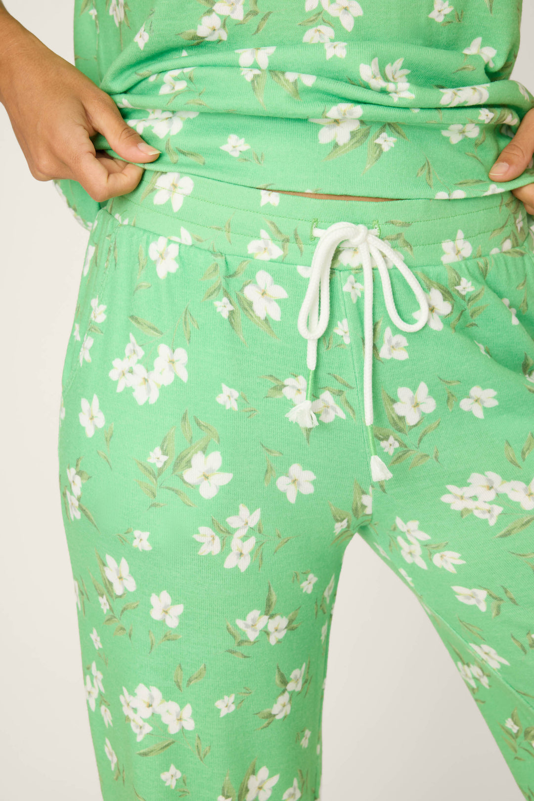 A person is shown adjusting the white drawstring waistband of P.J. Salvage's C'est La Vie Floral Print Open Leg Pants, featuring a green color and white floral pattern.
