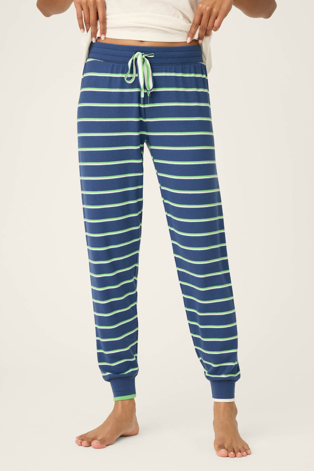 A person models the P.J. Salvage C'est La Vie Striped Banded Pants—blue jogger-style pajama bottoms featuring light green and white stripes with a matching drawstring—while standing barefoot against a plain background.