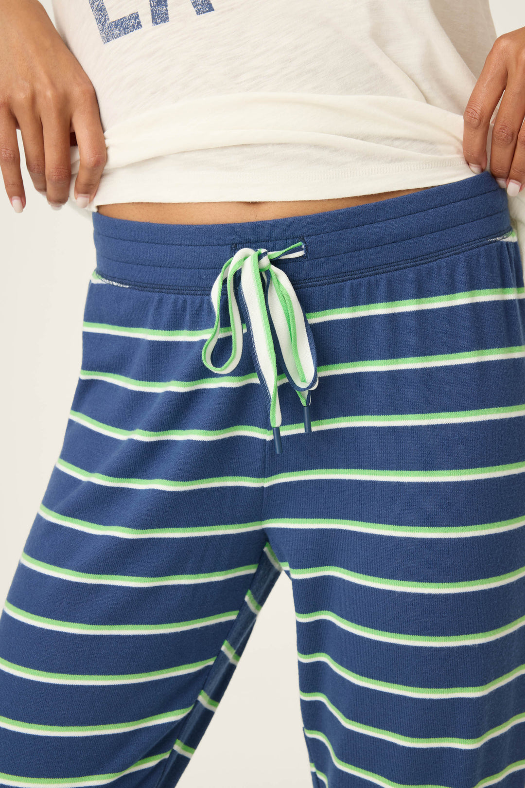 A close-up shows a person wearing P.J. Salvage's C'est La Vie Striped Banded Pants—blue with light green and white horizontal stripes, plus a matching drawstring at the waistband. The lower part of a white shirt is also visible.