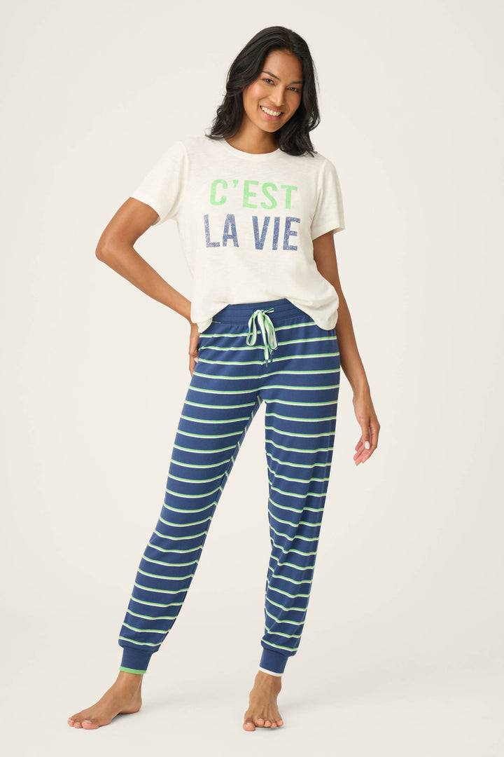 A woman smiles barefoot on a light background, wearing a white tee with "C’EST LA VIE" and P.J. Salvage's C'est La Vie Striped Banded Pants—blue with white and green stripes—her hand resting on her hip.