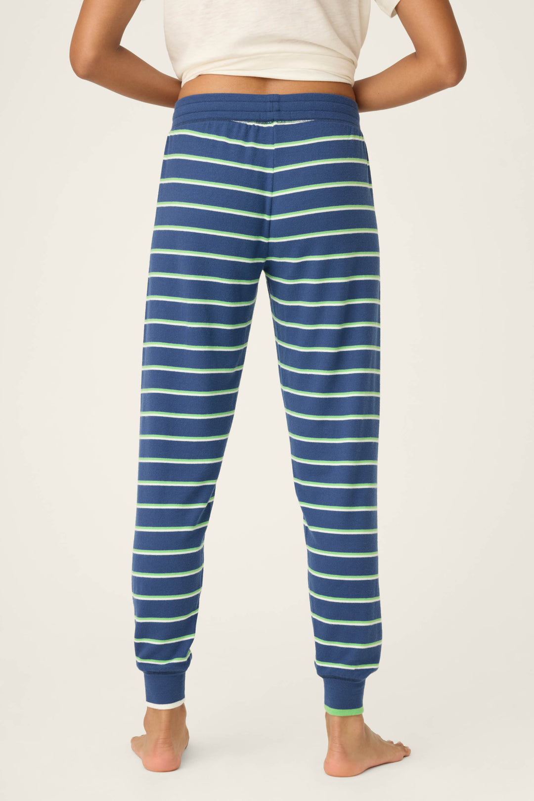 A person stands barefoot, back to the camera, wearing P.J. Salvage's C'est La Vie Striped Banded Pants—blue joggers with white and light green stripes—and a white T-shirt.
