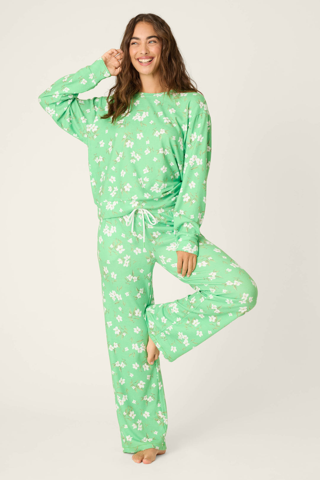 A person models the C'est La Vie Floral Print Lounge Set by P.J. Salvage, standing barefoot on one leg and smiling with a hand raised against a plain light background.