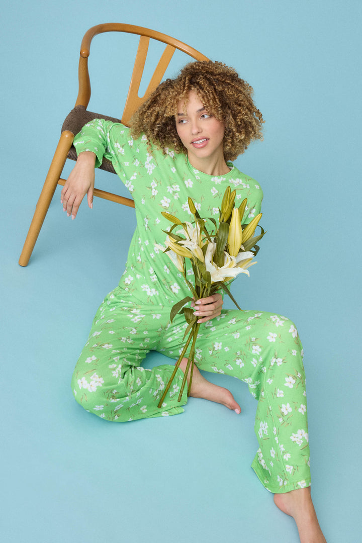 Wearing the P.J. Salvage C’est La Vie Floral Print Lounge Set, a person with curly hair sits on the floor against a wooden chair, holding white lilies, set against a light blue background.