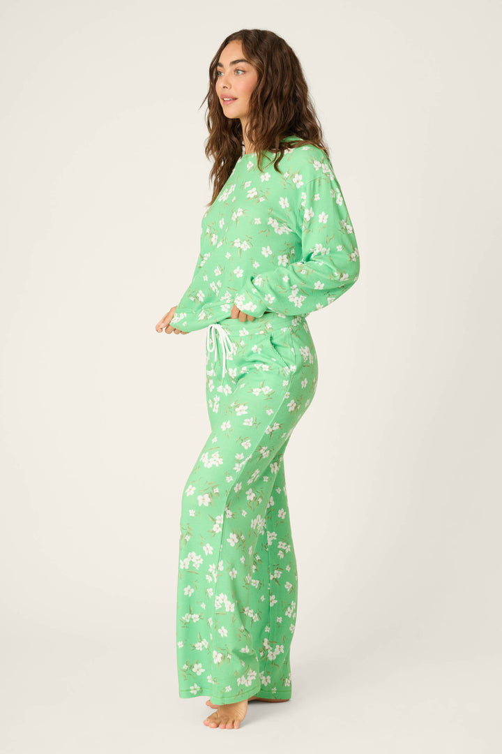 A person stands barefoot on a plain background, wearing the P.J. Salvage C'est La Vie Floral Print Lounge Set, featuring a matching green long-sleeve top and drawstring pants with a white floral pattern, facing sideways with hands in pockets.