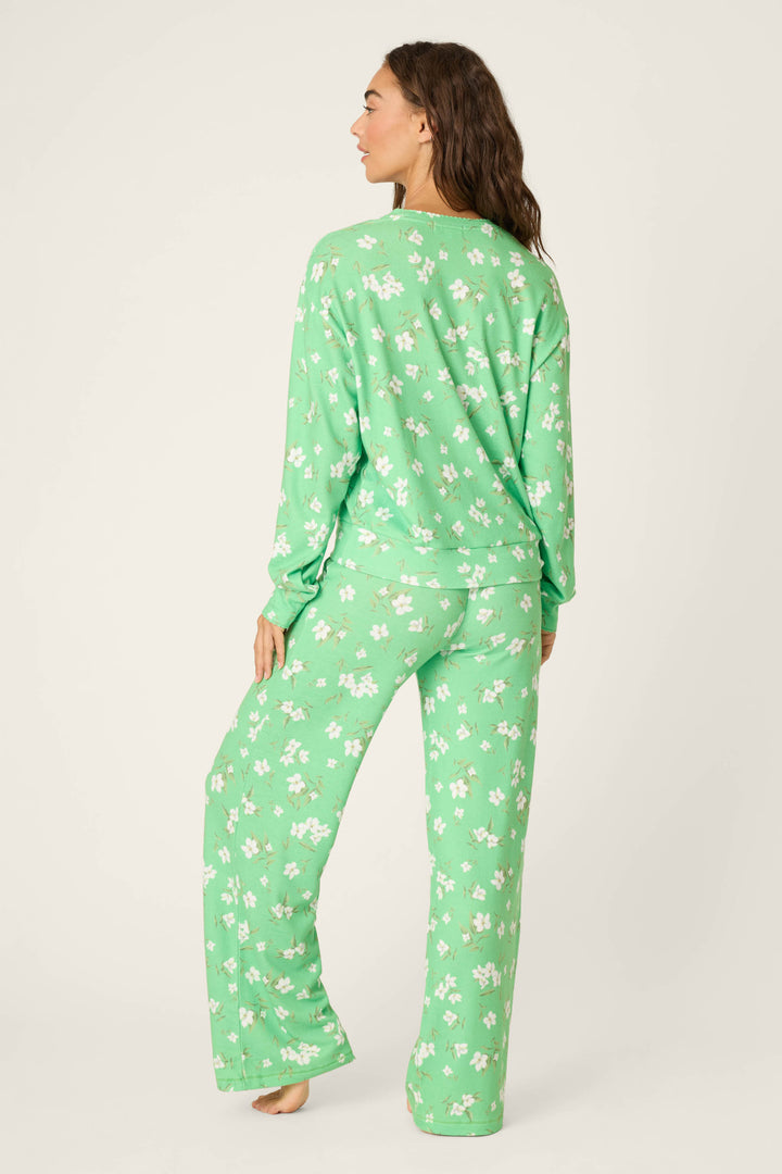 A person stands barefoot facing away, wearing the P.J. Salvage C'est La Vie Floral Print Lounge Set—loose-fitting, long-sleeve, green with a white floral design—against a plain light background.