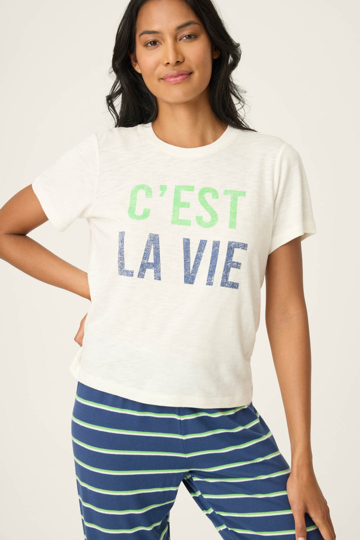 A person wears the P.J. Salvage C'est La Vie Graphic Print Short Sleeve Top, featuring green and blue text, paired with blue pants with green and white stripes, standing against a plain light background.