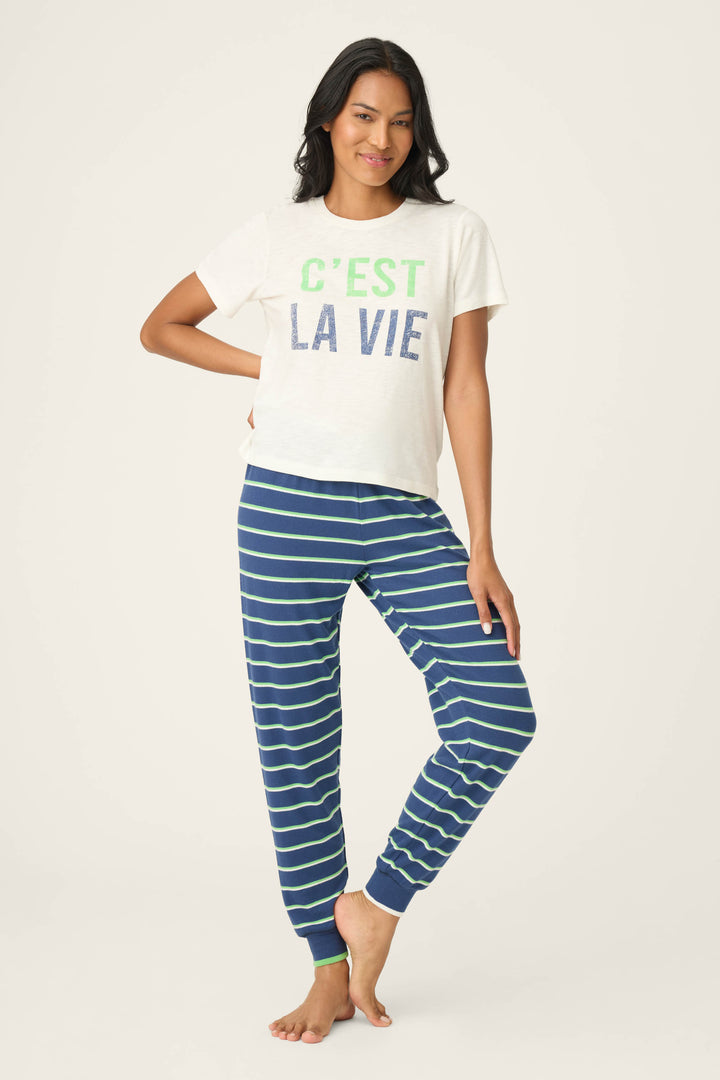 A woman, barefoot against a plain background, wears the P.J. Salvage C'est La Vie Graphic Print Short Sleeve Top and blue-and-green striped pajama pants. She poses with one hand on her hip and a slight smile.