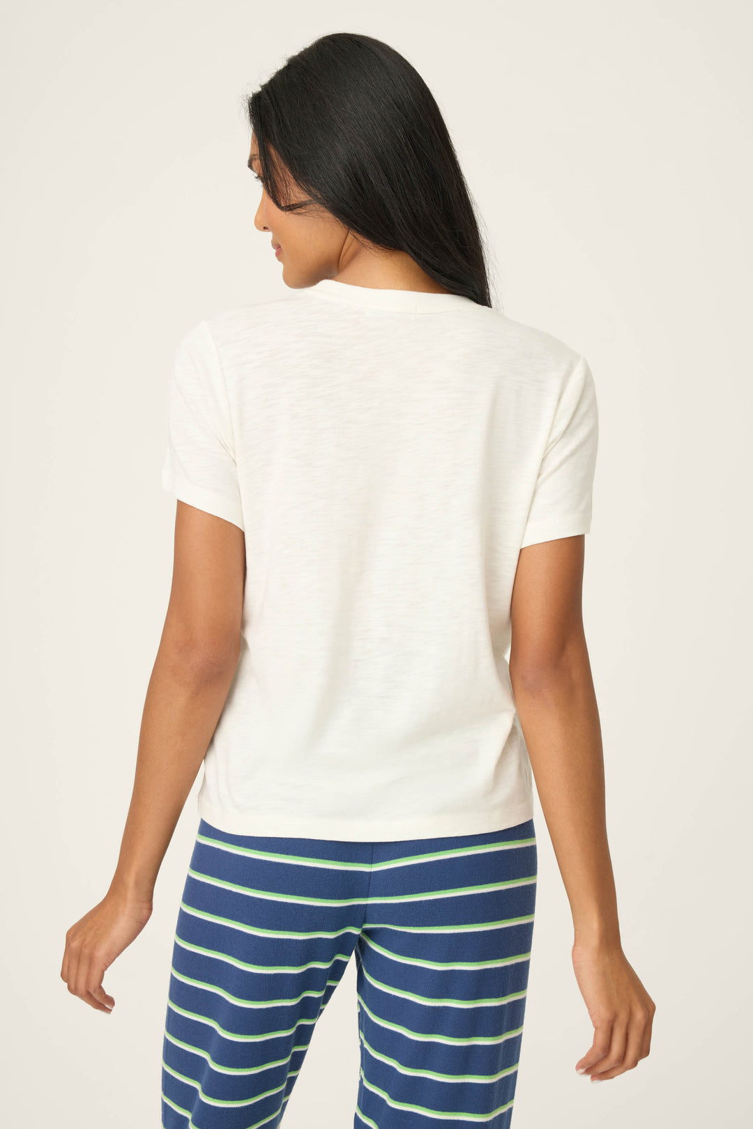 A person with long, dark hair is shown from the back, wearing the P.J. Salvage C'est La Vie Graphic Print Short Sleeve Top and blue pants with green and white horizontal stripes, standing against a neutral background.