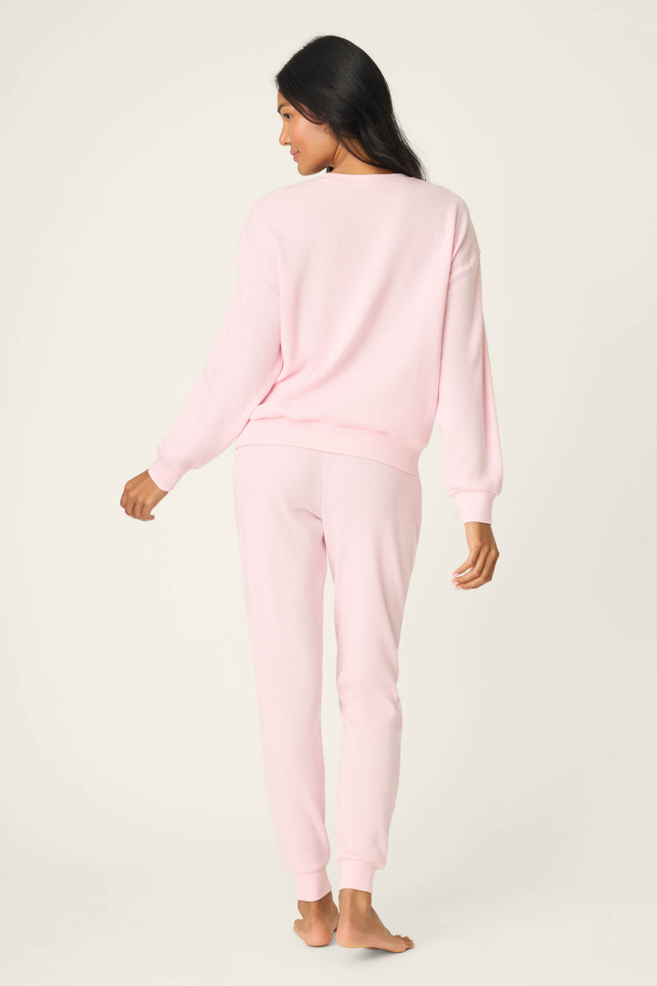 A person stands barefoot, back turned, in the P.J. Salvage Cross Stitch Casuals Ballet Pink Lounge Set—a soft, comfortable matching long-sleeve top and pants—against a plain, light-colored background.