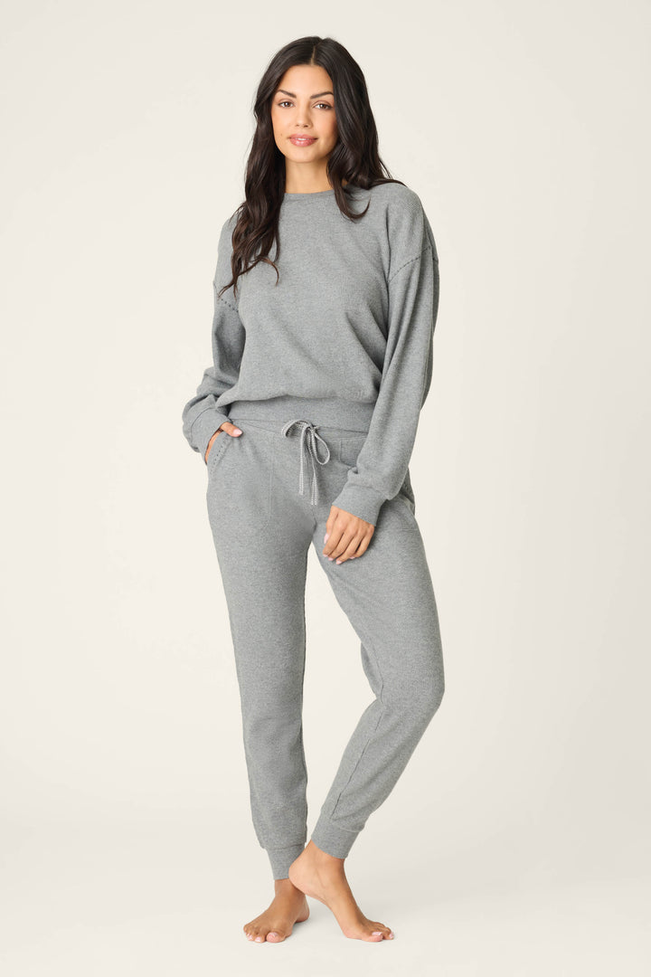 A person stands barefoot against a plain background, wearing the P.J. Salvage Cross Stitch Casuals Charcoal Lounge Set—featuring a matching long-sleeve sweatshirt and drawstring jogger pants—with one hand in a pocket and a relaxed expression.