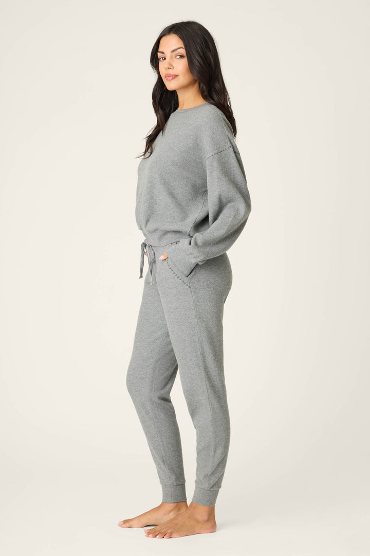 A woman stands barefoot on a plain background, wearing the P.J. Salvage Cross Stitch Casuals Charcoal Lounge Set—featuring a loose long-sleeve top and jogger sweatpants with pockets and a drawstring waist.