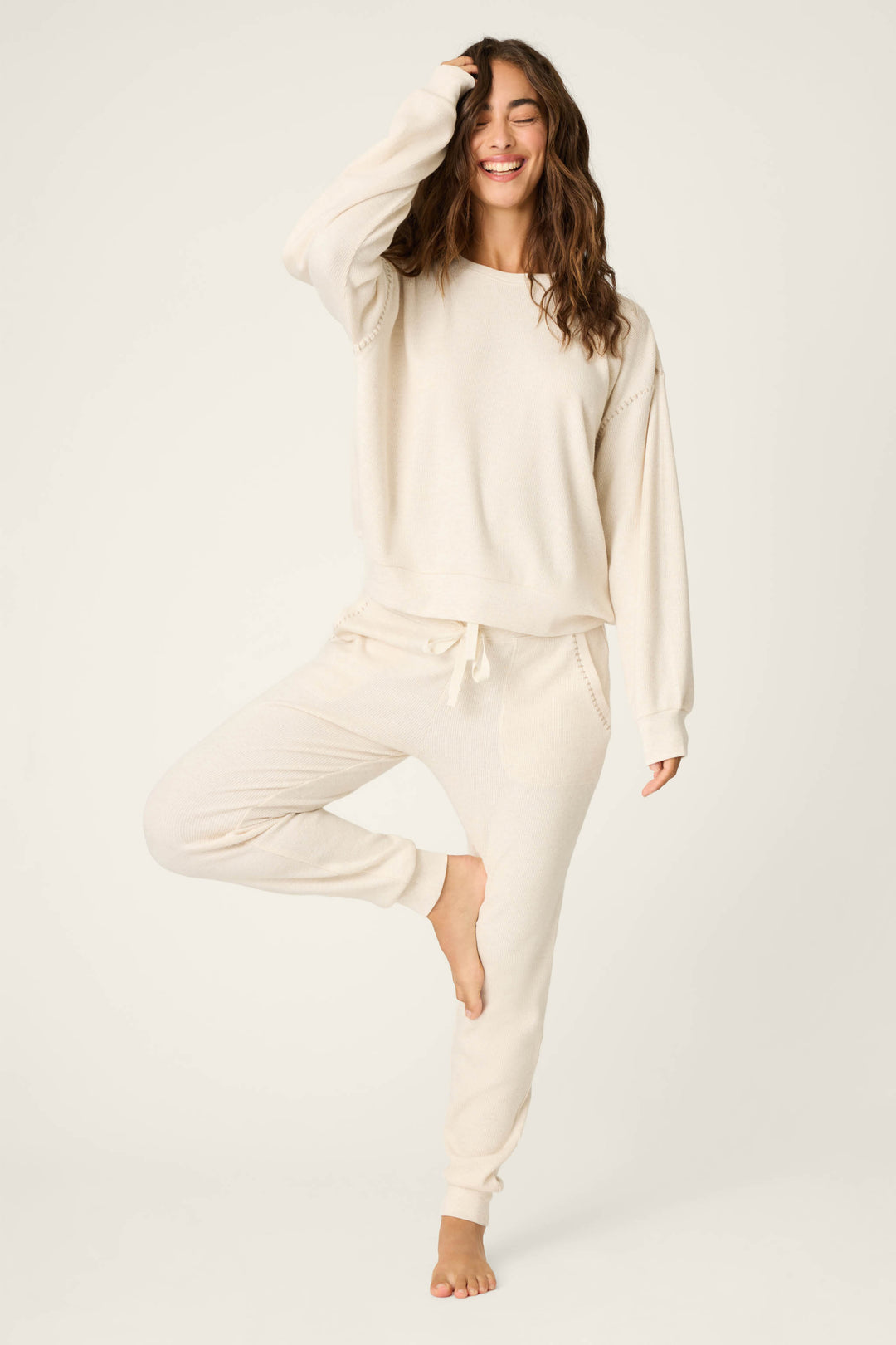 A person poses barefoot on one leg, smiling with a hand raised to their head, wearing the P.J. Salvage Cross Stitch Casuals Light Beige Lounge Set against a plain light background.