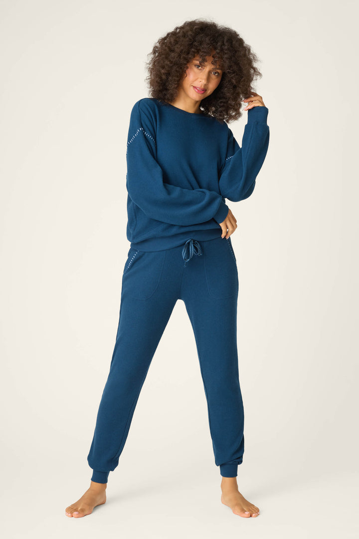 A person stands barefoot on a light background wearing the P.J. Salvage Cross Stitch Casuals Navy Lounge Set, with one hand touching their curly hair and the other resting on their hip.
