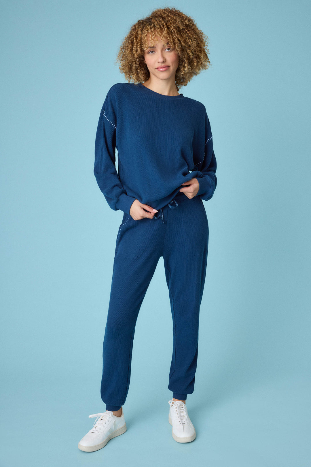 A person with curly hair stands against a light blue background, wearing the P.J. Salvage Cross Stitch Casuals Navy Lounge Set and white sneakers, with one hand in their pocket and the other on their hip.