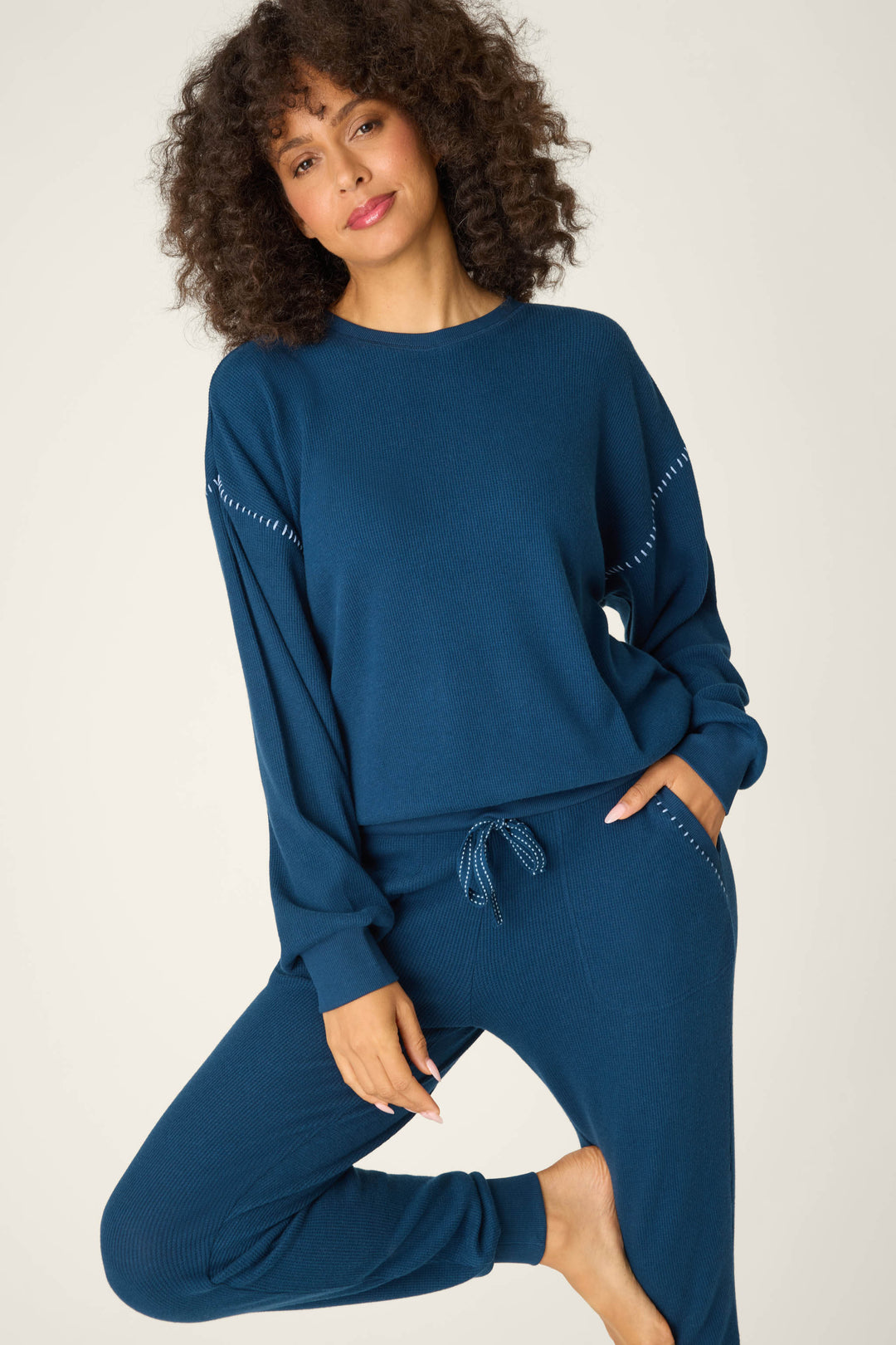 A person with curly hair wears the P.J. Salvage Cross Stitch Casuals Navy Lounge Set, standing on one leg against a plain light background with one hand in their pocket and a neutral expression.