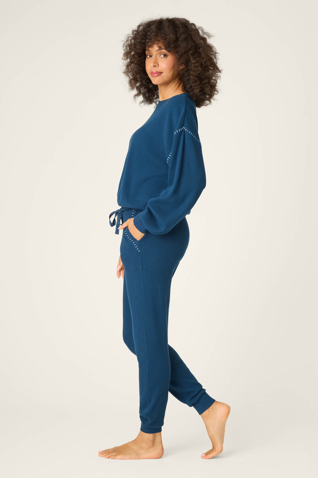A woman with curly hair stands barefoot on a plain background wearing the Cross Stitch Casuals Navy Lounge Set by P.J. Salvage, featuring matching dark blue knit sweater and jogger pants with visible stitching, looking at the camera with one hand in her pocket.