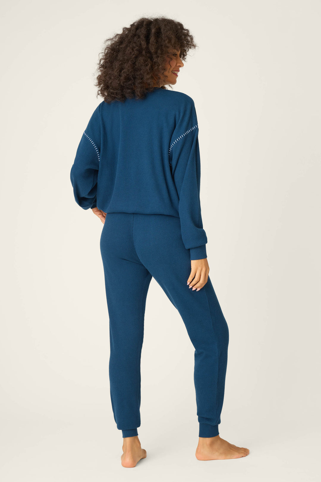 A person with curly hair stands barefoot on a light background, facing away, wearing the P.J. Salvage Cross Stitch Casuals Navy Lounge Set—featuring a matching, soft knit long-sleeve top and pants.