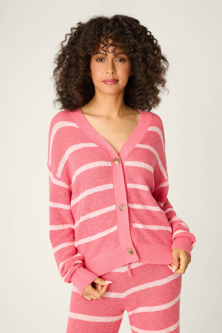 A person with curly hair models the P.J. Salvage Cozy Up Cotton Berry Kiss Cardigan, featuring pink and white stripes with brown buttons, paired with matching striped pants against a plain light background.