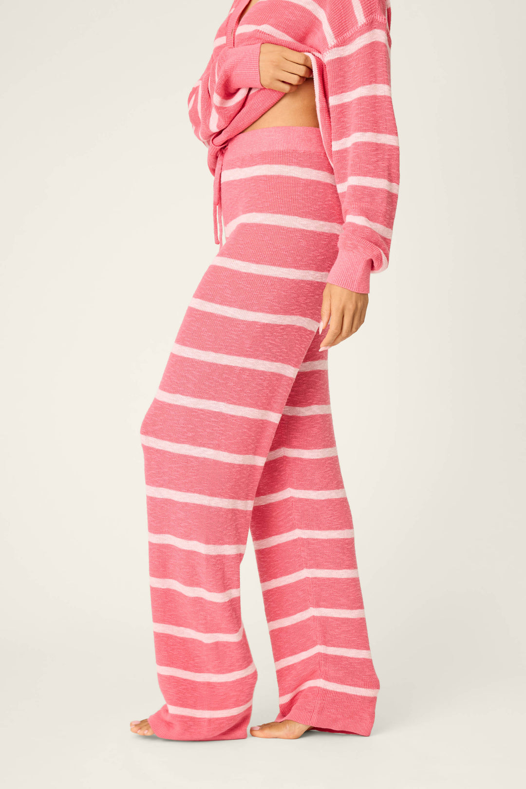 A person wears P.J. Salvage's Cozy Up Cotton Berry Kiss Open Leg Pants—loose-fitting, pink and white striped knit—with a matching top, barefoot on a neutral background, slightly lifting the top to reveal the pants' side.