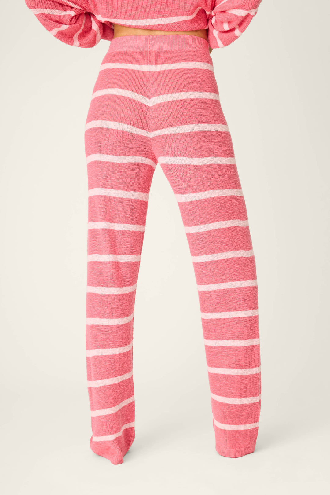 A person faces away from the camera wearing P.J. Salvage Cozy Up Cotton Berry Kiss Open Leg Pants, featuring a high waist, pink knit fabric with horizontal white stripes, a loose fit, and paired with a matching pink top.