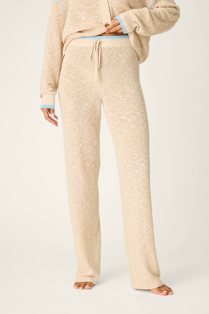 A person stands barefoot, wearing P.J. Salvage Cozy Up Cotton Heather Sand Open Leg Pants with a drawstring waist. The matching sweater has long sleeves and blue-accented cuffs. Only the lower body is shown.