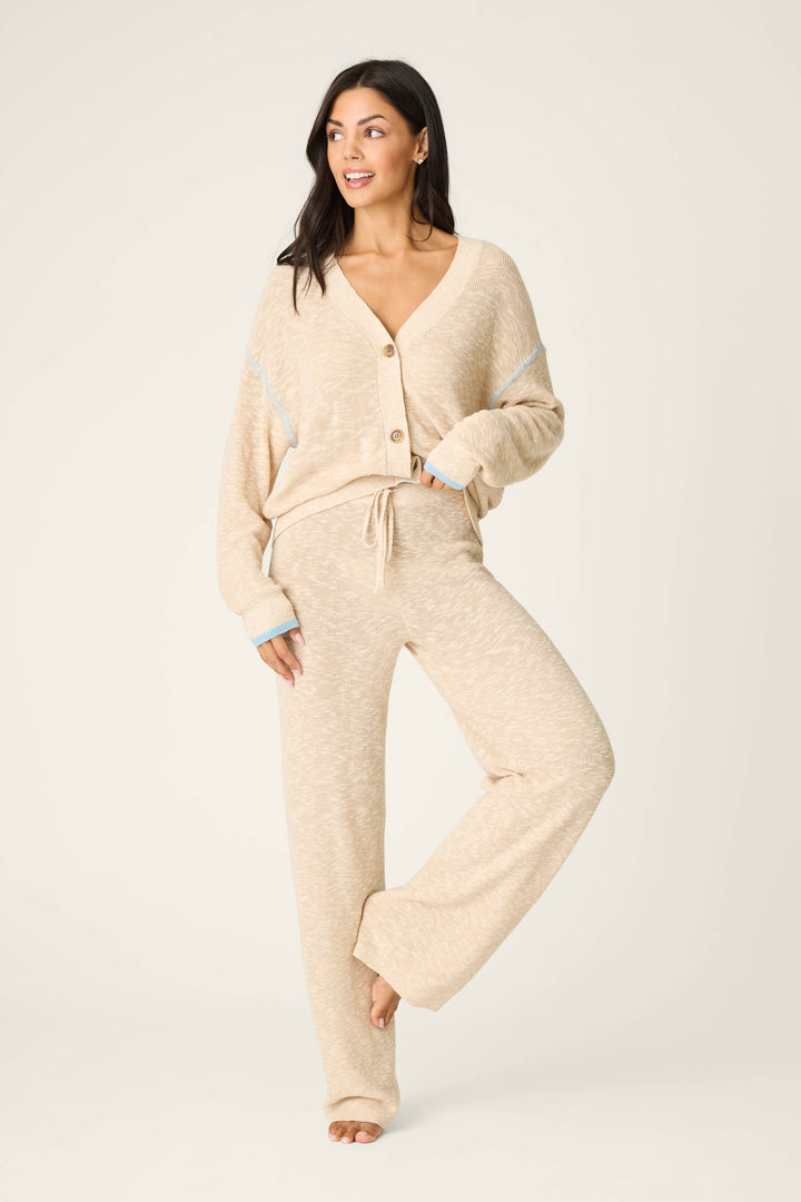 A woman stands on one leg, smiling to the side with a hand in her pocket, wearing P.J. Salvage Cozy Up Cotton Heather Sand Open Leg Pants and a loose knit cardigan against a plain light background.