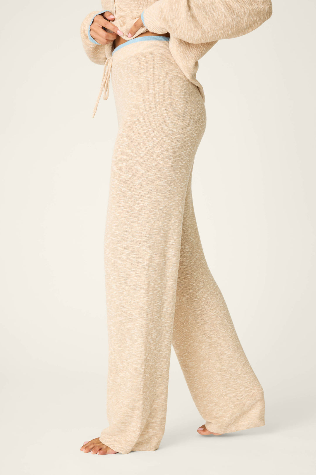 A person stands barefoot in P.J. Salvage Cozy Up Cotton Heather Sand Open Leg Pants, shown from the side, shoulders to feet, with hands adjusting the high-waisted waistband.