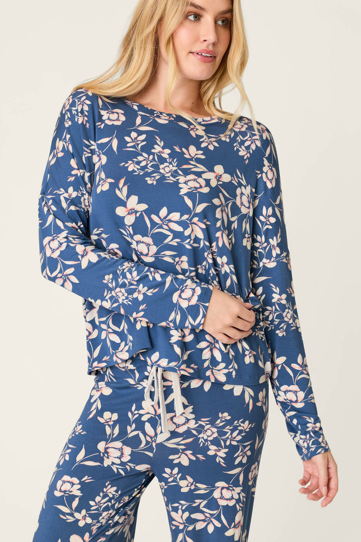 A woman wears the P.J. Salvage Flora Dreams Twilight Garden Long Sleeve PJ Top, featuring a blue background with a light floral print, paired with matching pants and standing against a plain light backdrop.