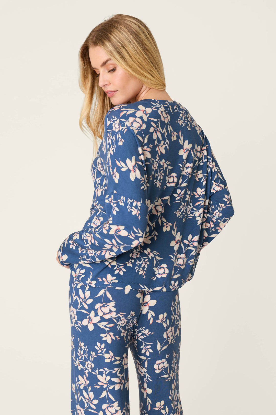 A woman with long blonde hair stands turned slightly away, wearing the P.J. Salvage Flora Dreams Twilight Garden Long Sleeve PJ Top, featuring a light pink and white floral print, against a plain light background.