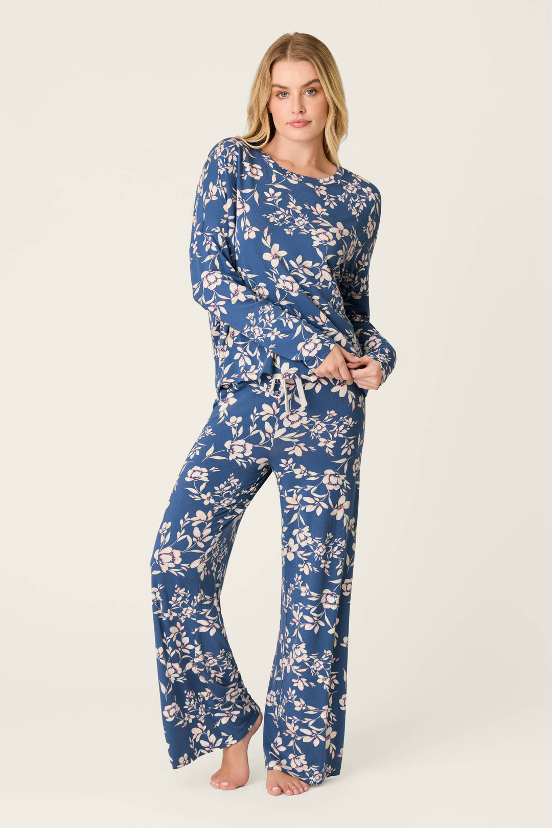 A woman stands barefoot in P.J. Salvage Flora Dreams Twilight Garden Open Leg PJ Pants, featuring soft modal jersey and a white and pink floral print, paired with a matching long-sleeve top against a plain light background.