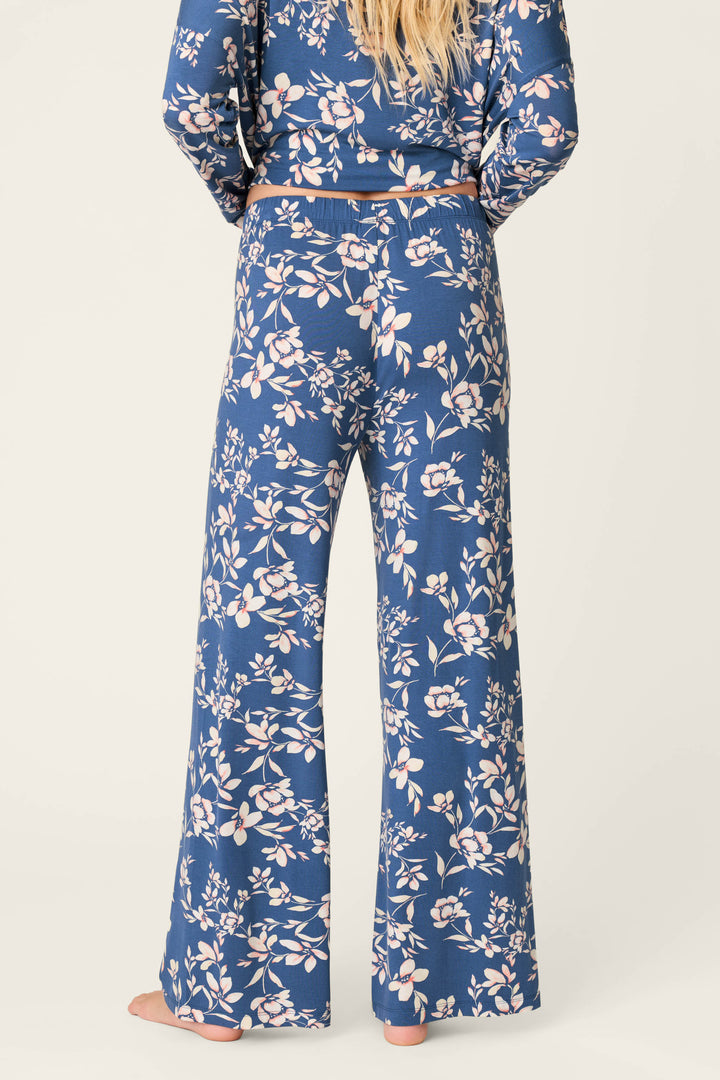 A person stands barefoot facing away, wearing P.J. Salvage Flora Dreams Twilight Garden Open Leg PJ Pants with a blue, white and pink floral pattern, paired with a matching long-sleeve top against a plain light background.