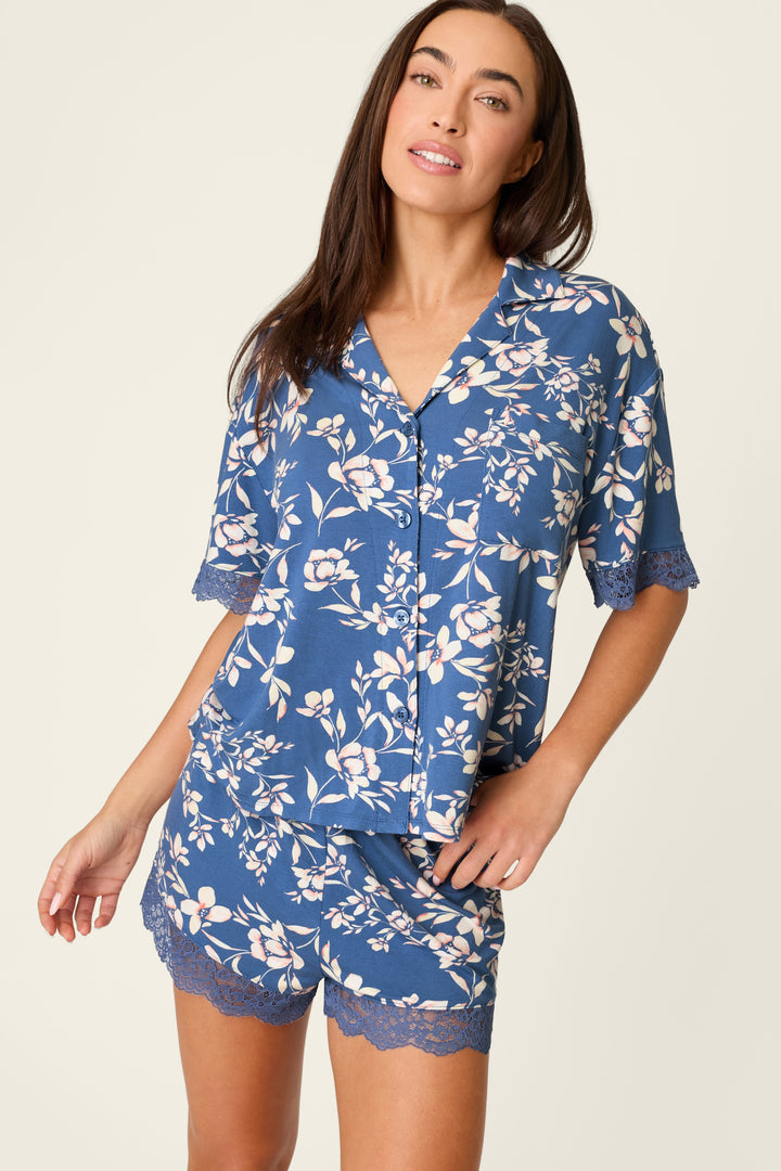 A woman smiles and poses with one hand on her hip, wearing the P.J. Salvage Flora Dreams Twilight Garden Short PJ Set, made from soft modal jersey with delicate lace trim, against a light background.