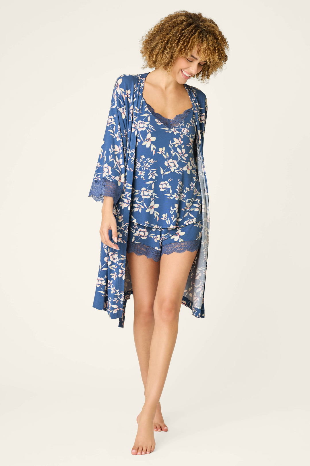 A woman with curly hair smiles barefoot, looking down, in a P.J. Salvage Flora Dreams Twilight Garden Robe, featuring blue floral print and soft modal jersey, against a plain light background.