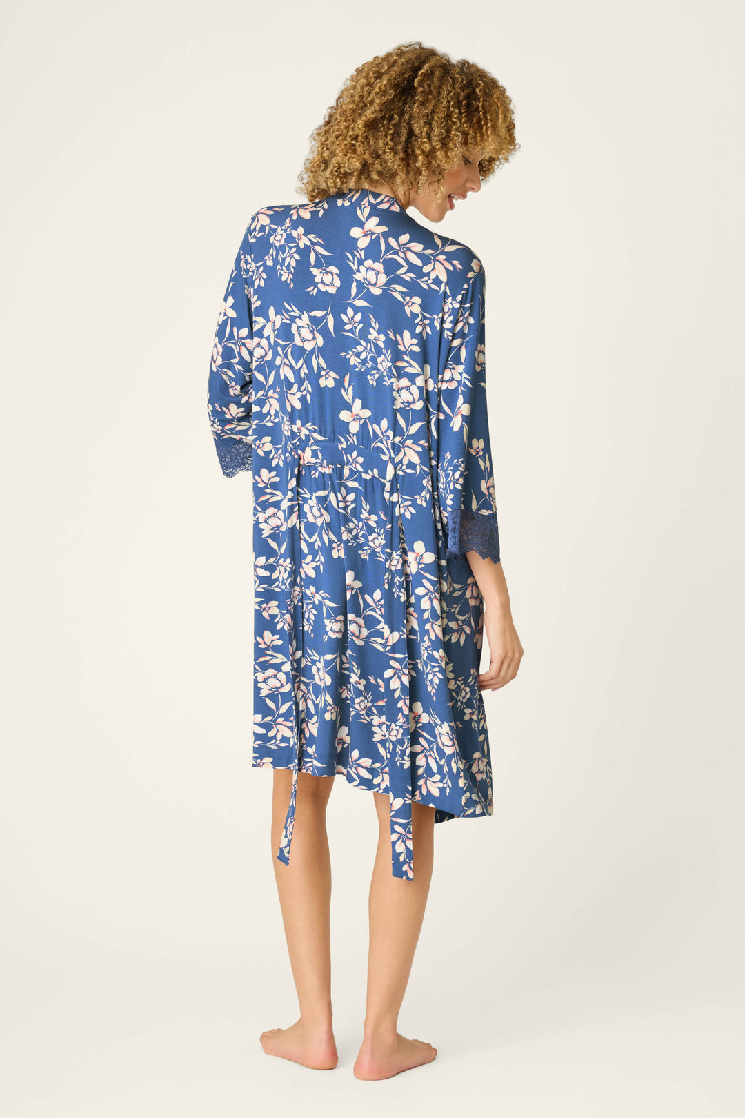 A person with curly hair stands barefoot facing away, wearing the P.J. Salvage Flora Dreams Twilight Garden Robe—a blue, knee-length floral modal jersey robe with white patterns, lace-trimmed sleeves, and a loosely hanging belt.