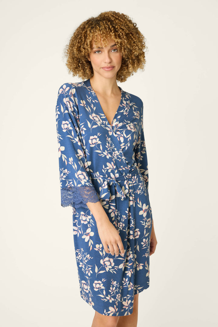 A person with curly hair, smiling gently at the camera, wears the P.J. Salvage Flora Dreams Twilight Garden Robe—blue with floral print and lace-trimmed sleeves—standing against a plain, light background.