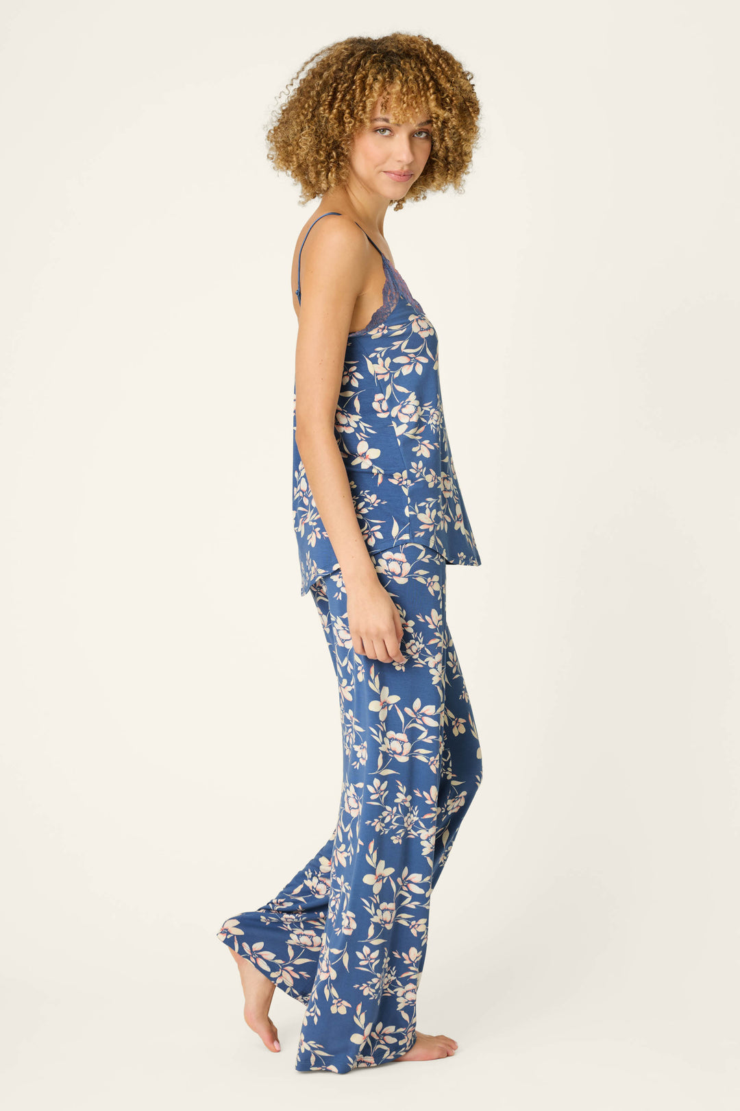 A woman with curly hair stands barefoot against a plain background, smiling softly at the camera while wearing the P.J. Salvage Flora Dreams Twilight Garden Long PJ Set—a silky modal jersey pajama set in a blue floral print.