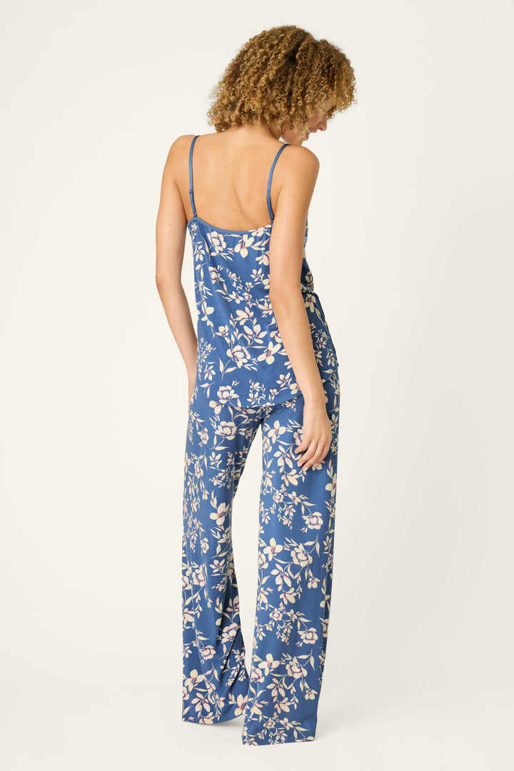 A woman stands facing away, wearing the Flora Dreams Twilight Garden Long PJ Set by P.J. Salvage—a blue floral sleep set crafted from silky modal jersey with thin straps and wide-leg pants. Her curly hair falls loose, and she has one hand in her pocket. The background is plain off-white.