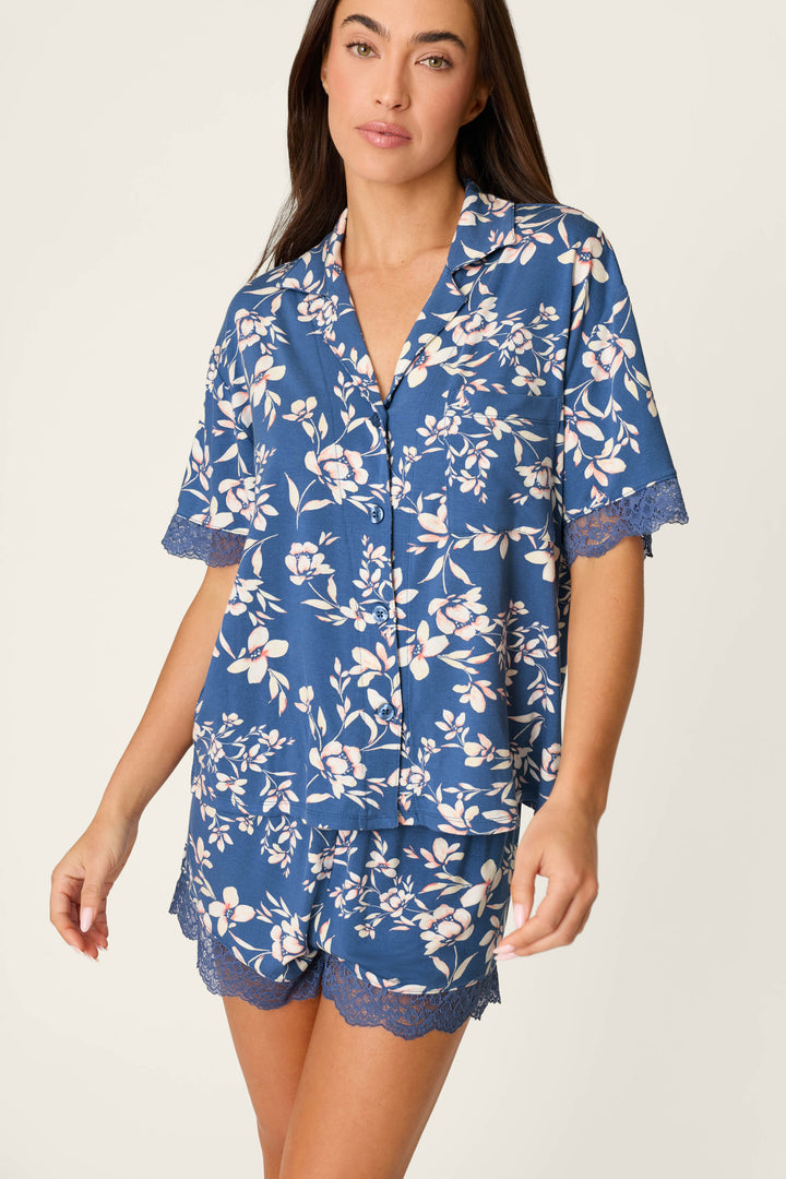 A woman wears the P.J. Salvage Flora Dreams Twilight Garden Short Sleeve Top, a relaxed-fit modal jersey pajama top with blue floral print and lace-trimmed collar, paired with matching shorts against a plain, light background.