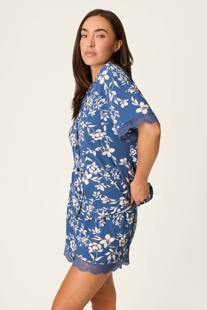 A woman with long brown hair confidently poses against a plain light background, hands in pockets, wearing the P.J. Salvage Flora Dreams Twilight Garden Short Sleeve Top in soft blue floral modal jersey.