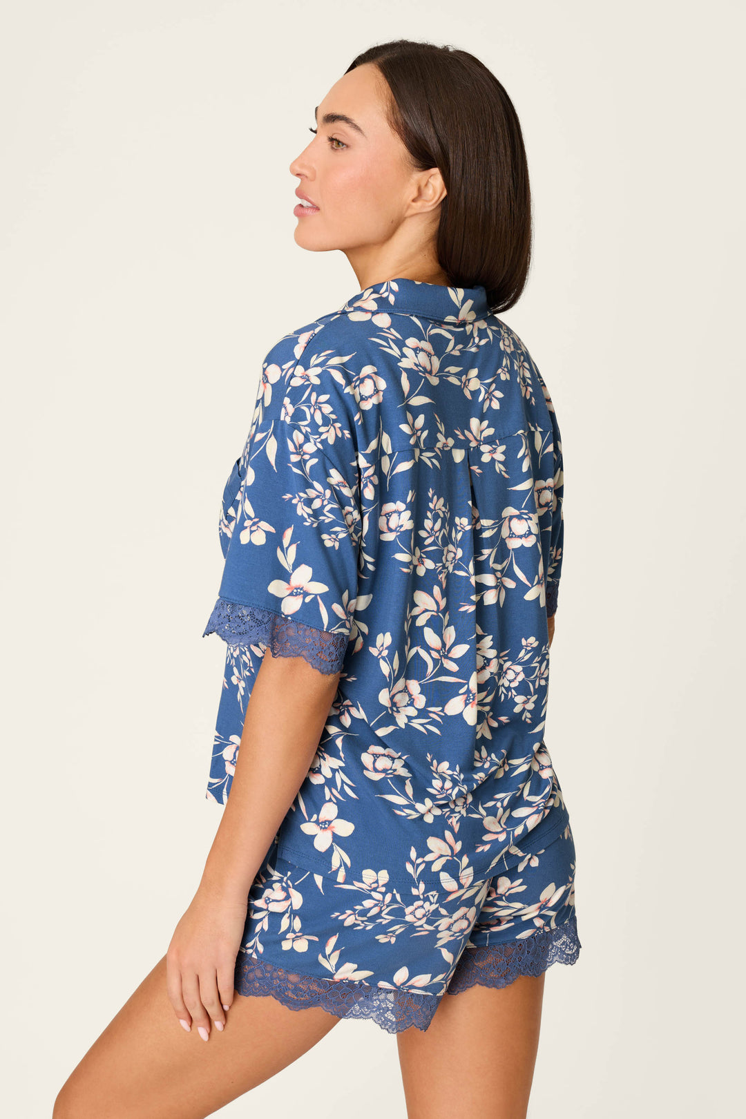 A woman with straight brown hair models the P.J. Salvage Flora Dreams Twilight Garden Short Sleeve Top, featuring blue floral print and lace trim in soft modal jersey, shown from the side and back against a plain light background.
