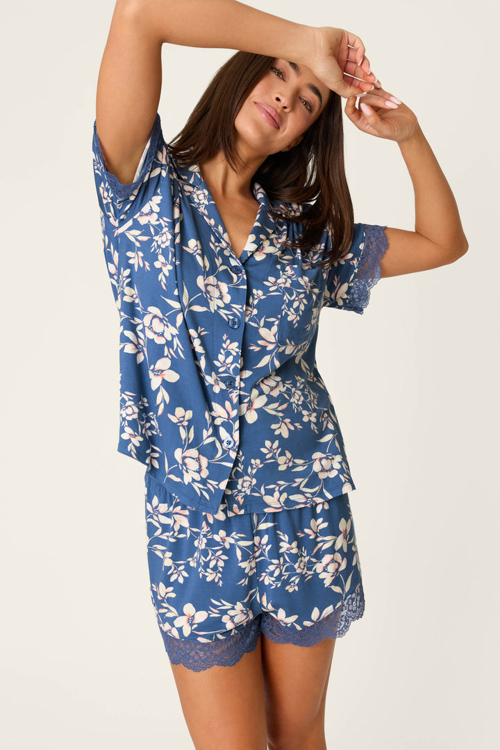 A woman models the P.J. Salvage Flora Dreams Twilight Garden Short Sleeve Top, a soft modal jersey pajama with lace trim, raising an arm and touching her forehead against a plain light background.
