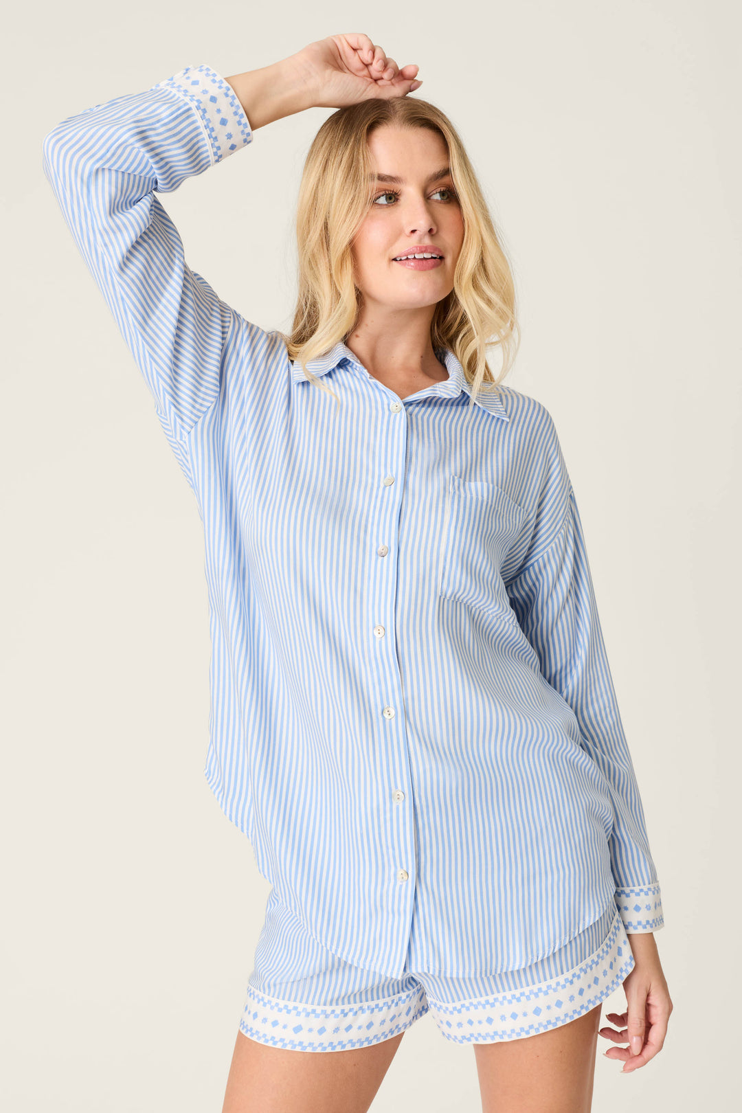 A woman stands against a plain background wearing the P.J. Salvage Frutta Fresca Striped Long Sleeve Button Up Top, raising one arm above her head and smiling slightly. This chic women's sleepwear top is both comfortable and stylish.