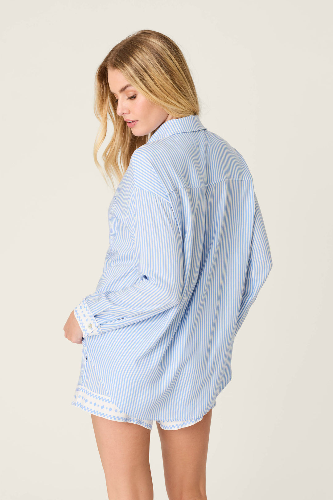A woman with long blonde hair faces away, modeling the P.J. Salvage Frutta Fresca Striped Long Sleeve Button Up Top—an oversized women’s sleepwear shirt with blue-and-white stripes and white lace trim—set against a plain light background.