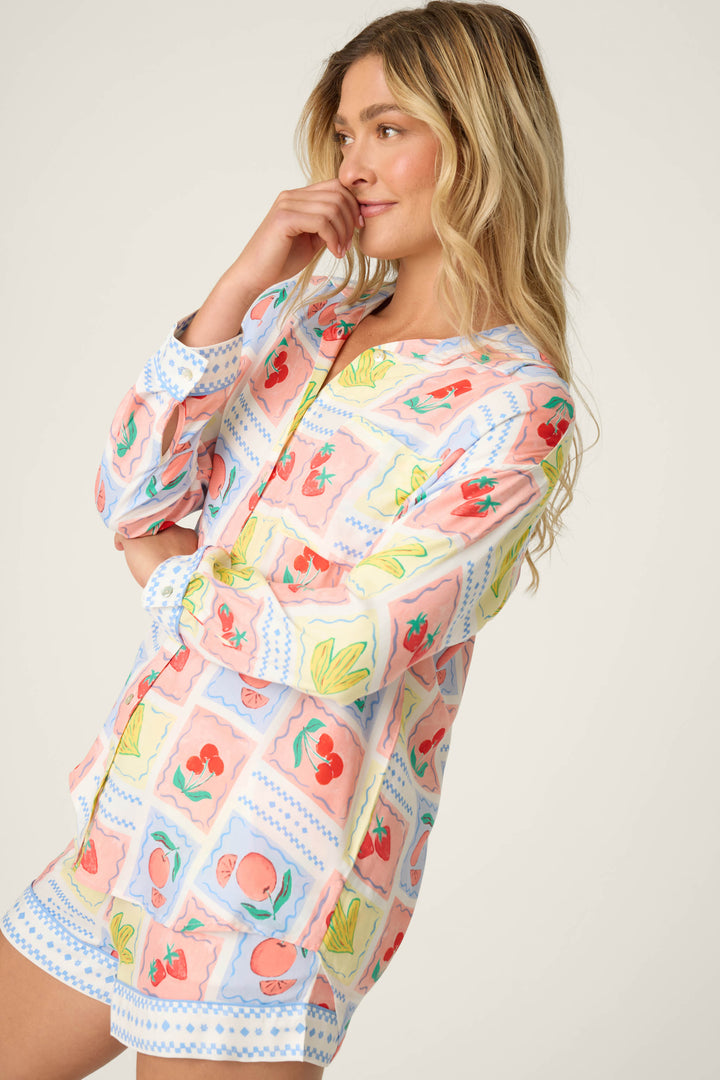 A person with long, wavy blonde hair wears the P.J. Salvage Frutta Fresca Summer Harvest Long Sleeve Button Up Top in a multicolor fruit print and stands sideways, one hand near their face.