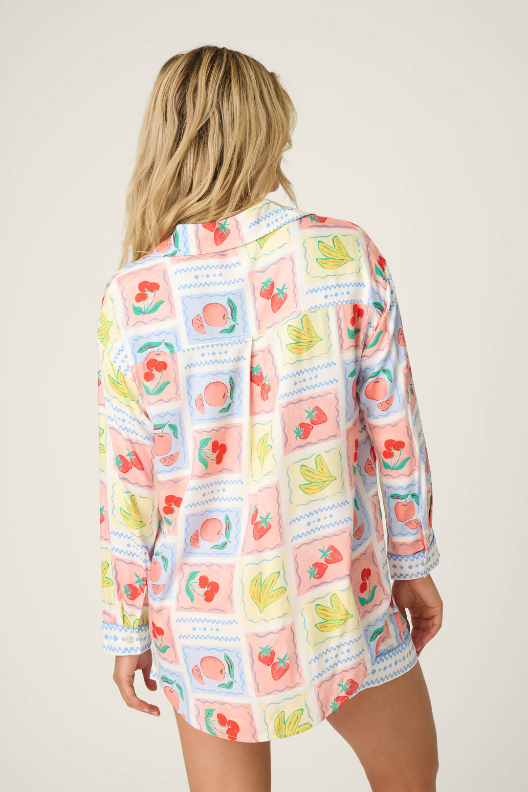 A person with long blonde hair is shown from the back, wearing the P.J. Salvage Frutta Fresca Summer Harvest Long Sleeve Button Up Top, a silky rayon sateen pajama shirt featuring a colorful fruit and floral square pattern on a light background.