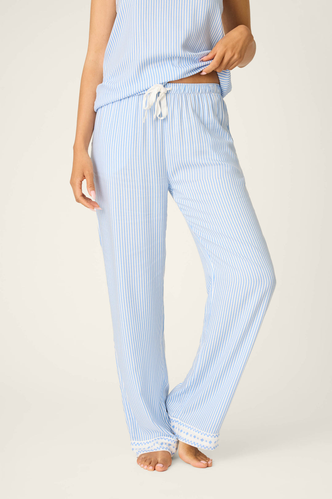 A person wears P.J. Salvage’s Frutta Fresca Striped Open Leg Pants in blue and white with a white drawstring and matching sleeveless top, barefoot against a plain light background for a cozy sleepwear look.