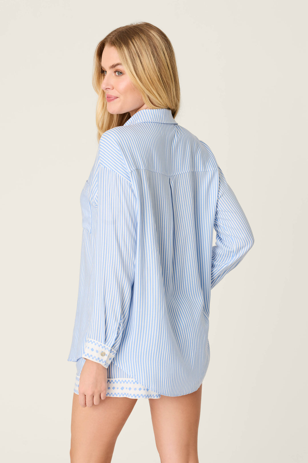 A woman with long blonde hair models the P.J. Salvage Frutta Fresca Striped Short Lounge Set in light blue and white, featuring patterned cuffs, as she looks over her shoulder against a plain background.