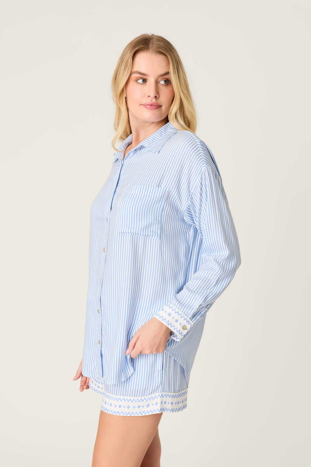 A woman with long blonde hair models the P.J. Salvage Frutta Fresca Striped Short Lounge Set, featuring light blue and white stripes with patterned trim at the cuffs and hem. She stands sideways, one hand in her pocket, gazing left.