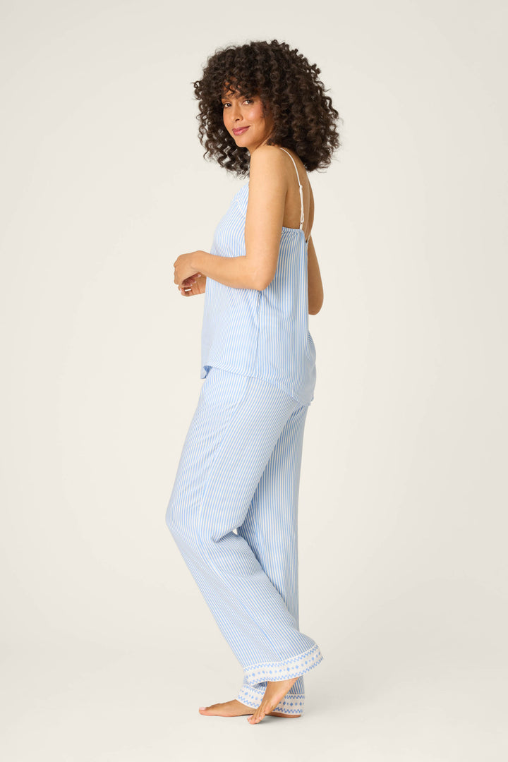 A woman with curly hair stands barefoot on a plain background, modeling the P.J. Salvage Frutta Fresca Striped Long Lounge Set with a sleeveless top and long pants, glancing over her shoulder.