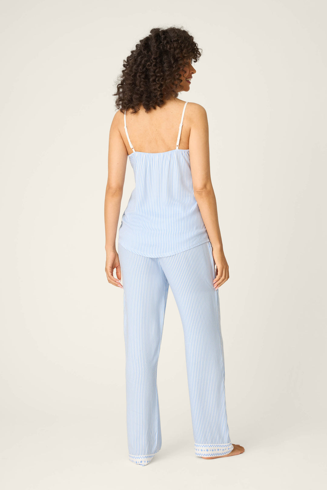 A woman stands barefoot facing away from the camera, wearing the P.J. Salvage Frutta Fresca Striped Long Lounge Set. Her shoulder-length curls and relaxed pose highlight the comfort of this blue-and-white sleepwear against a plain background.